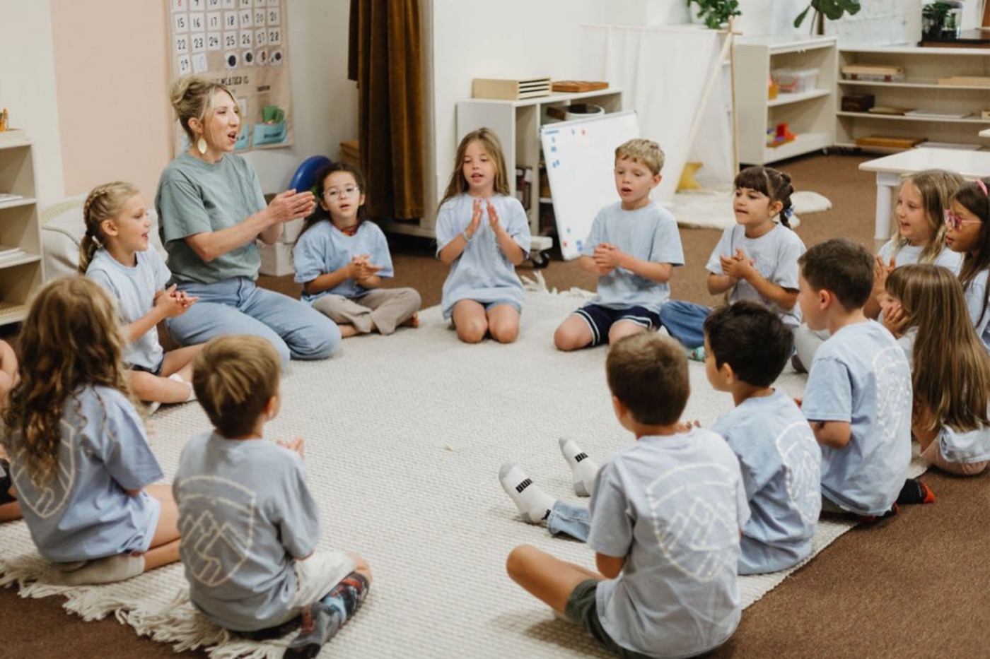 Teacher and kids in light-blue shirts seated in a circle on a rug, hands together, mid circle-time lesson