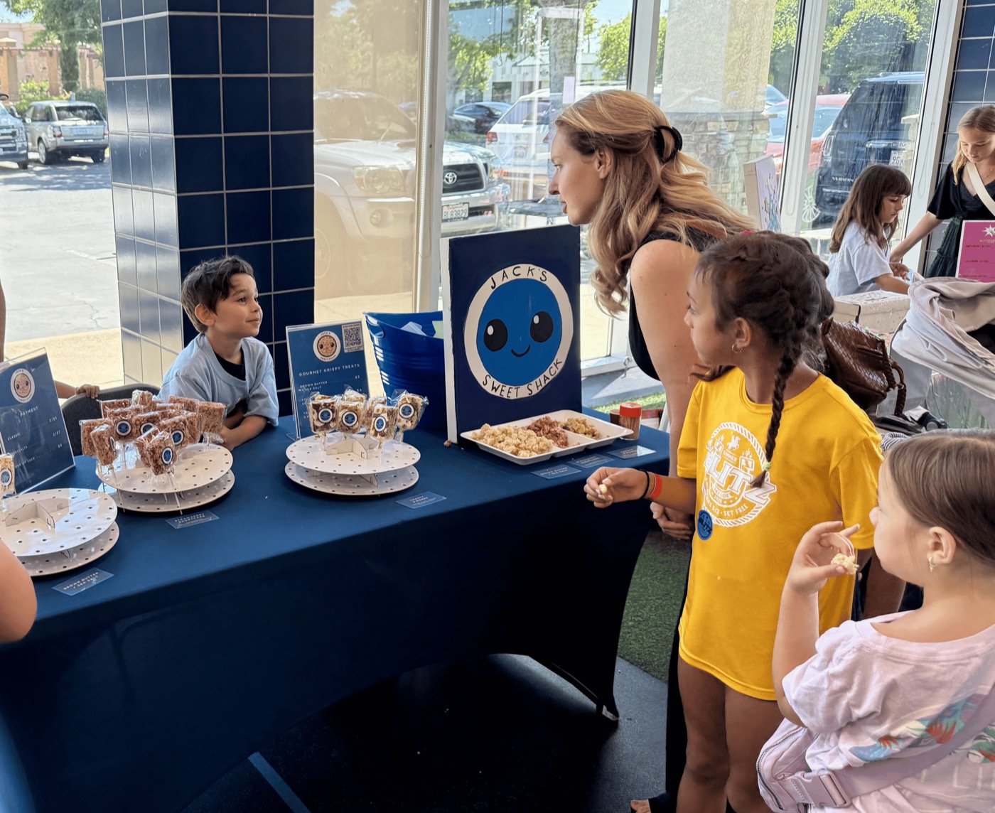 Student-run treat booth ('Jack's Sweet Shack') at a school showcase: a boy behind a navy table while kids and a parent browse